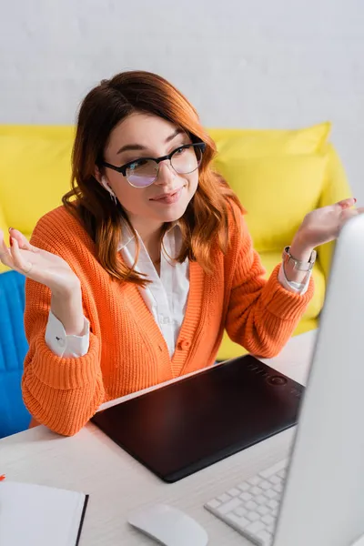 Donna positiva in auricolare mostrando gesto scrollata di spalle durante la videochiamata vicino tablet grafico e monitor sfocato — Foto stock