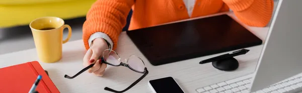 Vista parziale del ritoccatore che tiene gli occhiali vicino al tablet grafico, smartphone con schermo bianco e tazza da tè sulla scrivania, banner — Foto stock