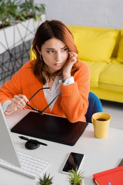 Diseñador gráfico cansado que sufre de dolor de cabeza mientras está sentado con gafas cerca de la tableta gráfica en el escritorio - foto de stock