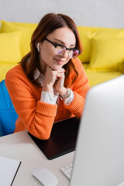 Glückliche Grafikdesignerin im Kopfhörer lächelt beim Videoanruf auf verschwommenem Computermonitor in der Nähe des Grafik-Tablets — Stockfoto