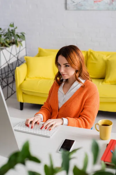 Bella donna digitando sulla tastiera vicino smartphone con schermo bianco e tazza di tè sulla scrivania — Foto stock