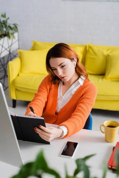 Woman in earphone writing in notebook near smartphone with blank screen and tea cup on desk — Stock Photo