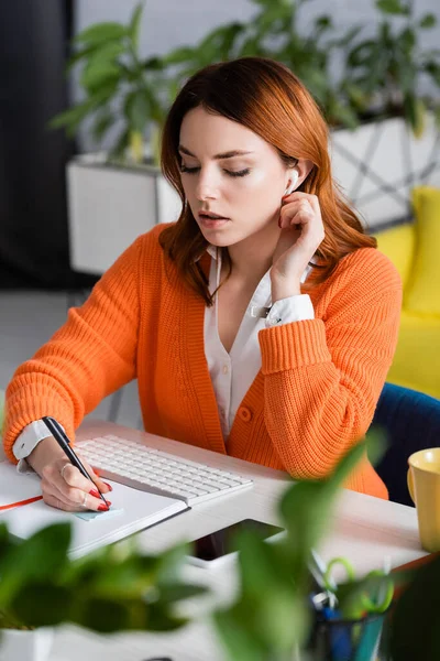 Woman in earphone writing in notebook near keyboard and smartphone on blurred foreground — Stock Photo
