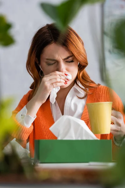 Mujer enferma con los ojos cerrados estornudos en servilleta de papel mientras sostiene la taza de té caliente en primer plano borroso - foto de stock