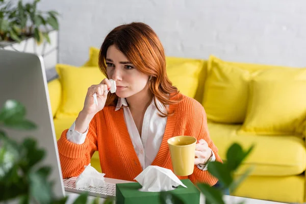 Mujer enferma con servilleta de papel y taza de té mirando el monitor de la computadora mientras trabaja en primer plano borroso - foto de stock