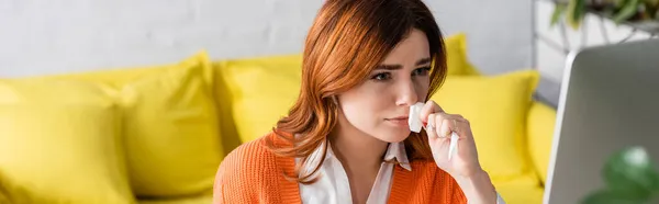 Kranke Frau mit Papierserviette, die in der Nähe eines verschwommenen Computermonitors arbeitet, Banner — Stockfoto