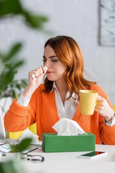 Sick woman with closed eyes sneezing in paper napkin while sitting at home workplace with cup of tea — Stock Photo