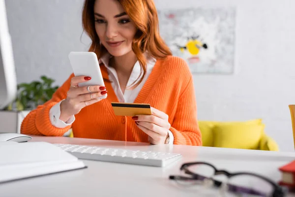 Verschwommene Frau lächelt, während sie ihre Kreditkarte in der Nähe der Tastatur zu Hause hält — Stockfoto