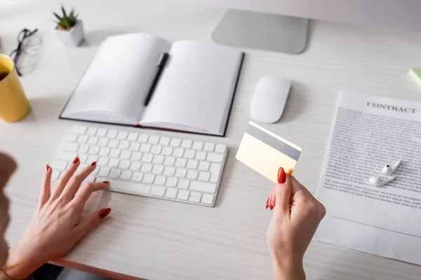 Vista recortada de la mujer escribiendo en el teclado mientras sostiene la tarjeta de crédito cerca del cuaderno en blanco en el escritorio - foto de stock