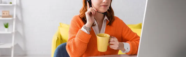 Vista recortada de la mujer en auriculares sosteniendo taza de té mientras trabaja cerca del monitor de la computadora, pancarta - foto de stock