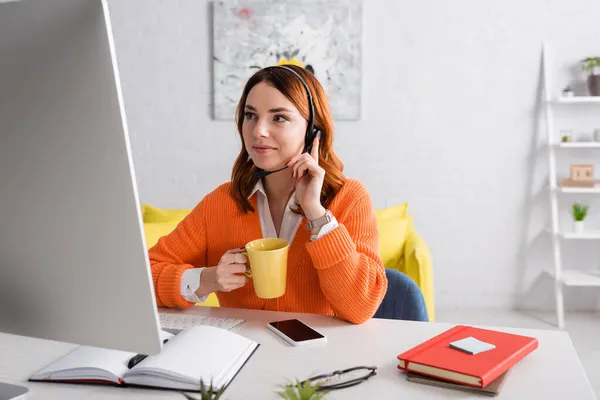 Mujer sonriente en auriculares sosteniendo la taza de té mientras trabaja cerca de la computadora y el teléfono inteligente con pantalla en blanco - foto de stock