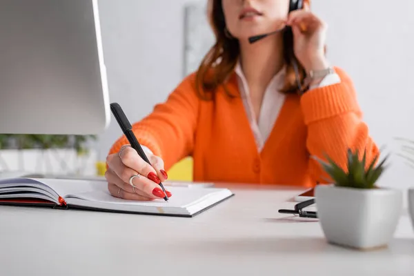 Vista recortada de la mujer borrosa en la escritura de auriculares en el cuaderno mientras trabaja en casa - foto de stock