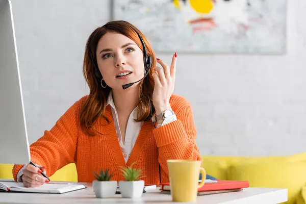 Hübsche junge Frau schreibt in Notizbuch, während sie zu Hause im Headset arbeitet — Stockfoto