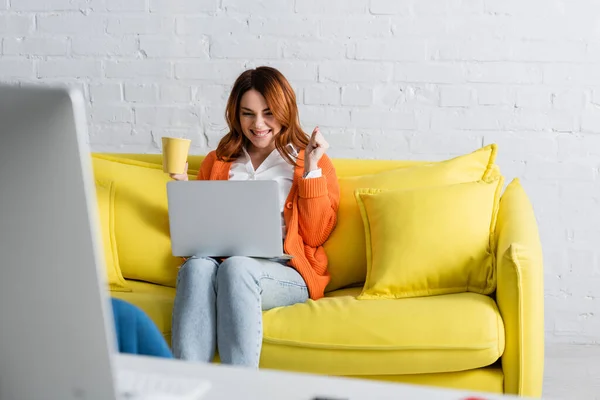 Mulher animada com xícara de chá trabalhando no laptop enquanto sentado no sofá perto do monitor do computador em primeiro plano desfocado — Fotografia de Stock