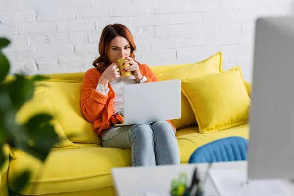 Young woman sitting on sofa with laptop and drinking tea on blurred foreground — Stock Photo
