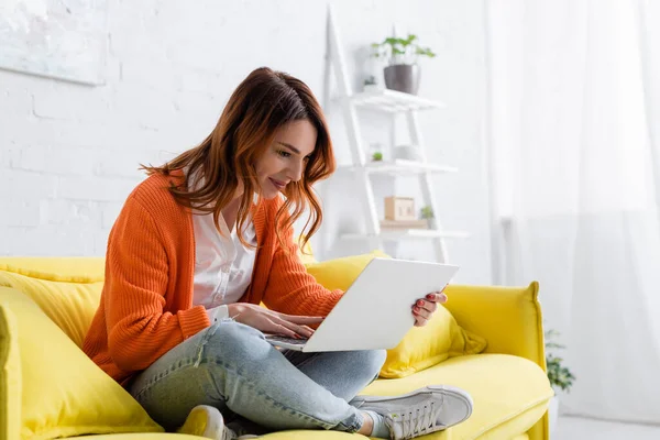 Cheerful freelancer sitting on yellow sofa with crossed legs and working on laptop — Stock Photo