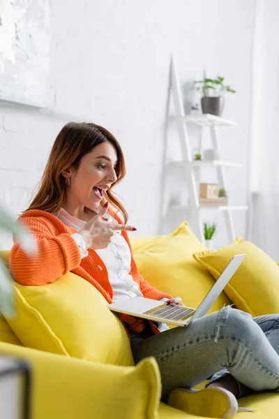 Amazed freelancer pointing at laptop while sitting on yellow couch on blurred foreground — Stock Photo