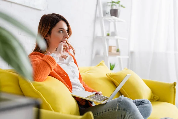 Nachdenklicher Freiberufler schaut weg, während er auf gelbem Sofa mit Laptop sitzt — Stockfoto