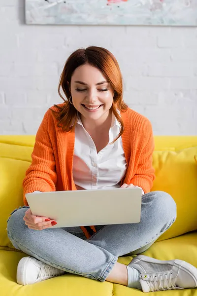 Allegro freelance che lavora su laptop seduto su un divano giallo con gambe incrociate — Foto stock