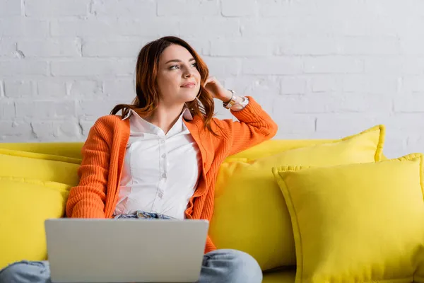 Verträumte und lächelnde Frau, die mit Laptop auf gelbem Sofa sitzt und wegschaut — Stockfoto