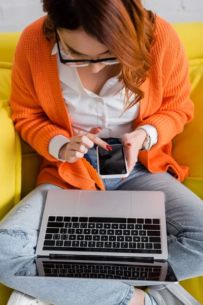 Vista de ángulo alto de la mujer en gafas de vista sentado en el sofá con el ordenador portátil y el uso de teléfono inteligente con pantalla en blanco - foto de stock