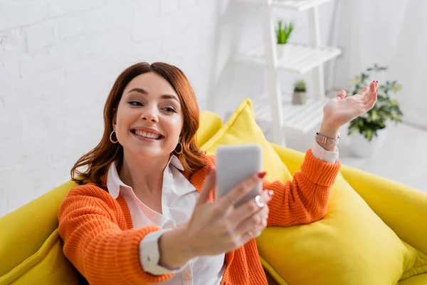 Happy young woman sitting on yellow sofa and taking selfie on mobile phone — Stock Photo