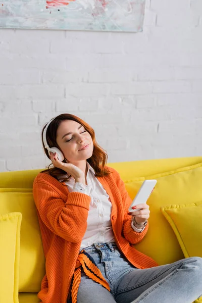 Femme souriante dans les écouteurs tenant smartphone tout en écoutant de la musique avec les yeux fermés — Photo de stock