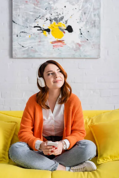 Happy woman with smartphone sitting on yellow couch and listening music in headphones — Stock Photo