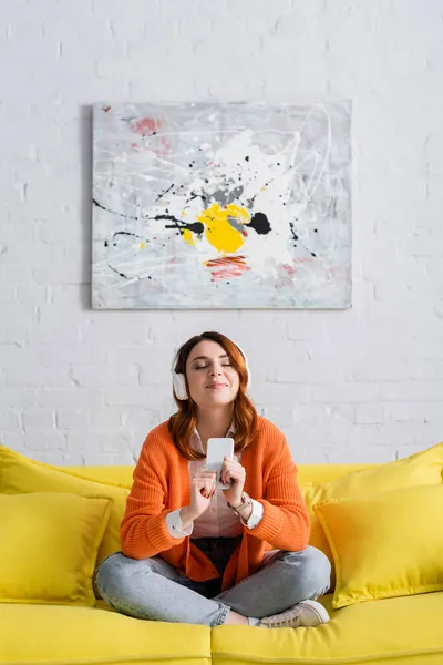 Femme heureuse avec les yeux fermés écouter de la musique tout en étant assis sur le canapé jaune avec les jambes croisées — Photo de stock