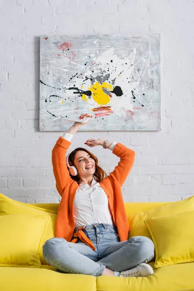 Fröhliche Frau, die mit erhobenen Händen auf dem Sofa sitzt und über Kopfhörer Musik hört — Stockfoto