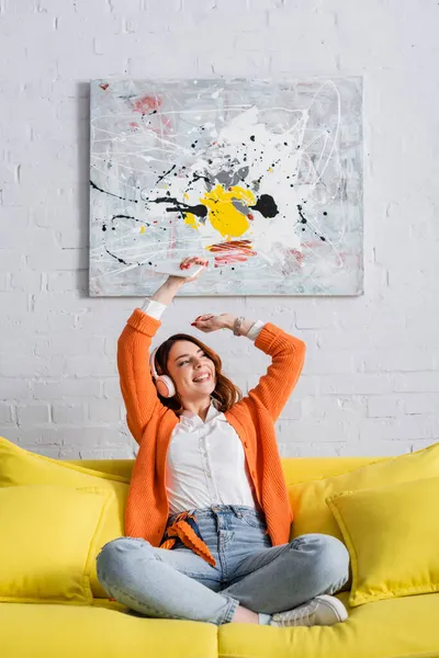 Femme joyeuse assise sur le canapé avec les mains levées tout en écoutant de la musique dans les écouteurs — Photo de stock