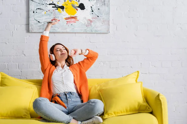 Excited woman with smartphone in raised hand listening music on yellow couch — Stock Photo