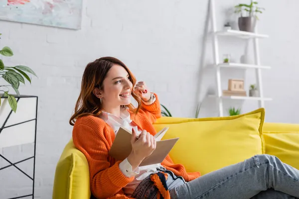 Glückliche Frau mit Buch schaut weg, während sie zu Hause auf dem Sofa sitzt — Stockfoto