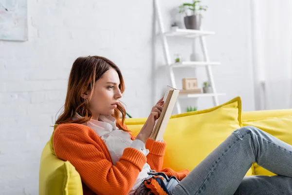 Livre de lecture femme tendue tout en étant assis sur canapé jaune — Photo de stock