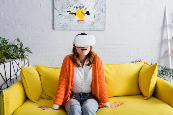 Erstaunt Frau in vr Headset sitzt auf gelbem Sofa im Wohnzimmer — Stockfoto