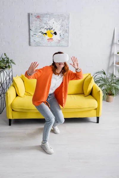 Vista completa de juego de mujer joven en auriculares vr cerca de sofá amarillo en la sala de estar - foto de stock