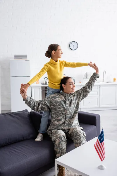 Mujer en uniforme militar cogida de la mano de su hija cerca de la bandera americana en casa - foto de stock