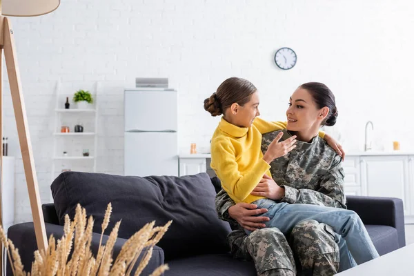 Femme souriante en uniforme de camouflage étreignant sa fille sur le canapé à la maison — Photo de stock
