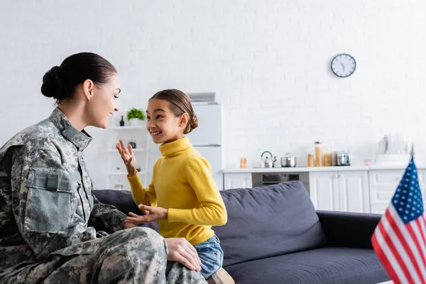 Smiling kid talking to mother in military uniform near blurred american flag at home — Stock Photo