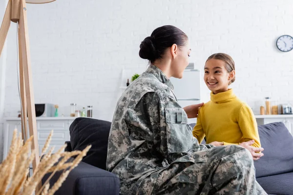 Mujer en uniforme de camuflaje mirando a su hija en el sofá en casa - foto de stock