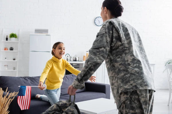 Criança sorridente com bandeira americana olhando para mãe desfocada em uniforme militar em casa — Fotografia de Stock