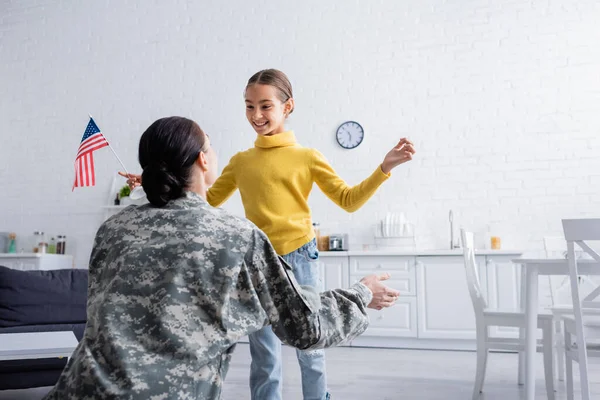 Cheerful kid holding american flag near parent in military uniform at home — Stock Photo