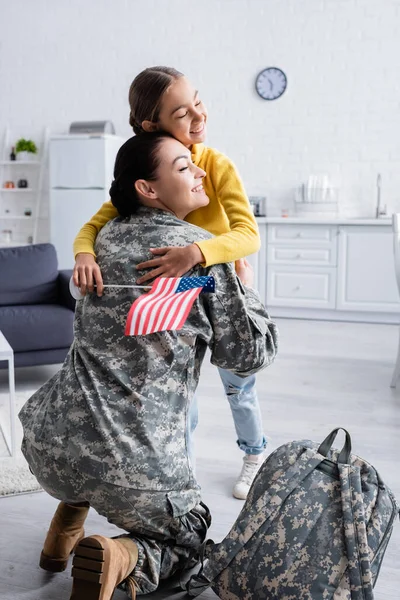 Criança com olhos fechados e bandeira americana abraçando a mãe em uniforme militar perto de mochila em casa — Fotografia de Stock