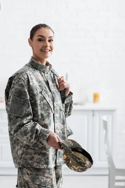 Soldado sonriente en uniforme militar sosteniendo mochila y gorra en casa - foto de stock