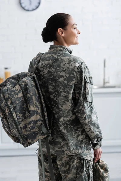 Vista lateral del soldado sonriente con la mochila sosteniendo la gorra en casa - foto de stock