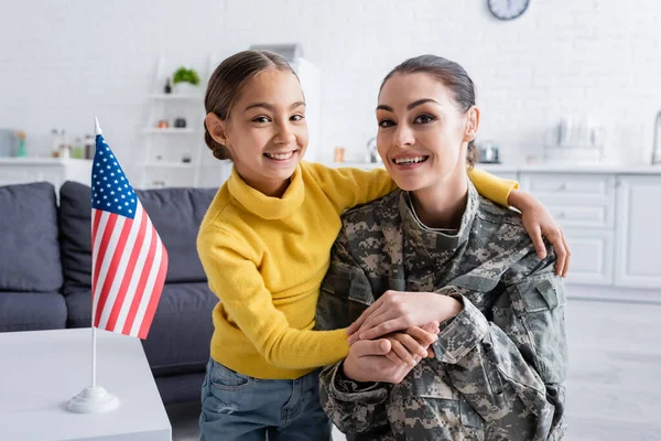 Mulher sorridente em uniforme militar segurando a mão da filha perto da bandeira americana em casa — Fotografia de Stock