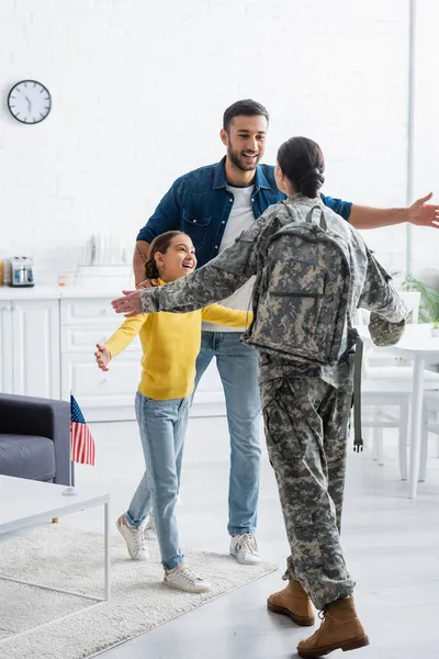 Feliz hombre y niño de pie cerca de la madre en uniforme militar y bandera americana en casa - foto de stock