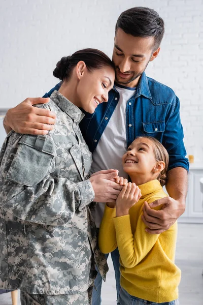 Uomo abbracciare moglie in uniforme camuffamento e figlia a casa — Foto stock