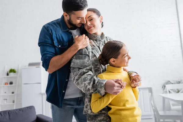 Sonriente niño cogido de la mano de la madre en uniforme militar cerca de papá en casa - foto de stock