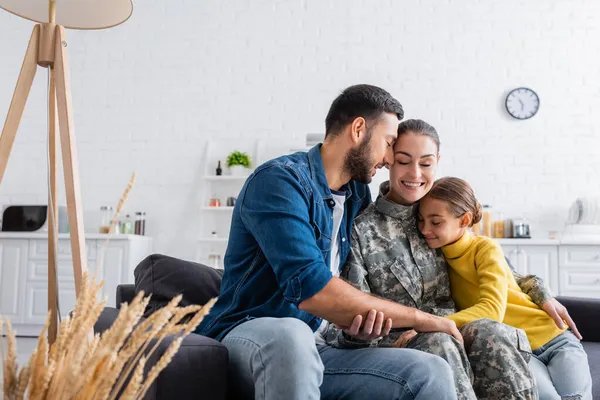 Soldato sorridente in uniforme seduto vicino a bambino e marito sul divano a casa — Foto stock